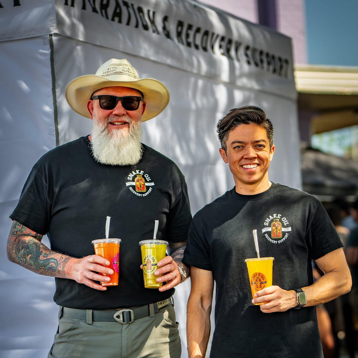 Jeff and John wearing black staff t-shirts, holding ready-to-drink Snake Oil in front of a white tent at a street fair.
