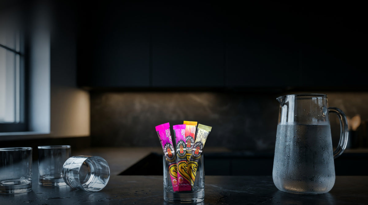 Snake Oil stick packs in a rocks glass on a dark kitchen counter beside a pitcher of water and empty glasses, early morning