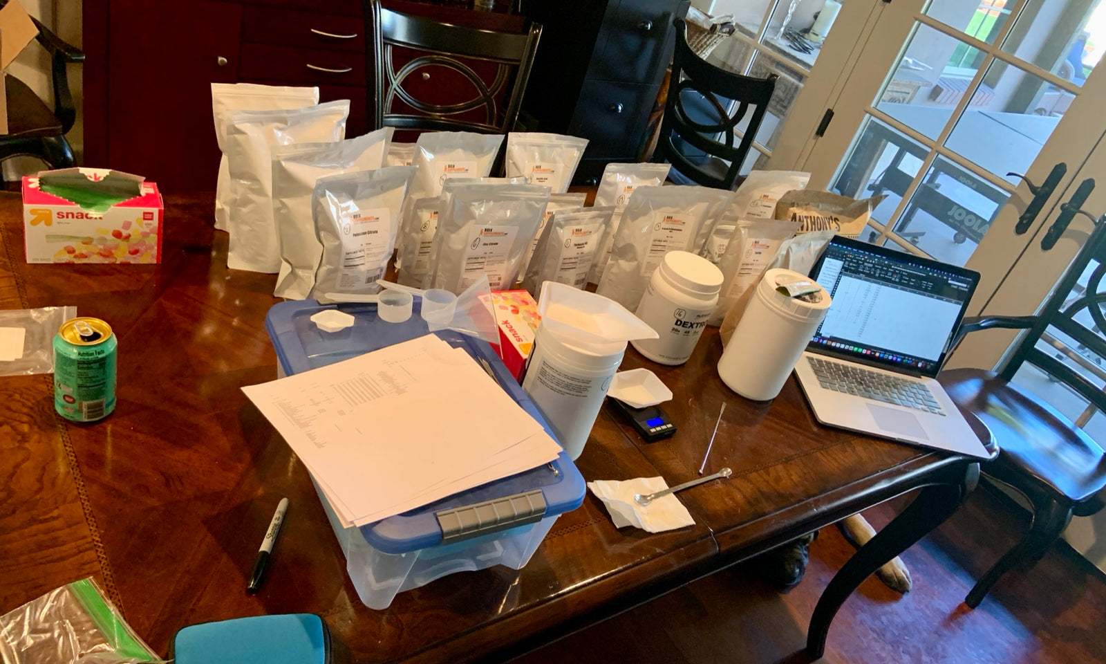 Ingredients, a kitchen scale, and handwritten recipe notes spread across a home dining table during early formulation